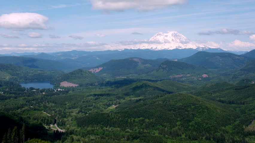 Snow-capped Mount Rainier towers above rolling forested hills and a scenic lake under a partly cloudy sky in Washington State.