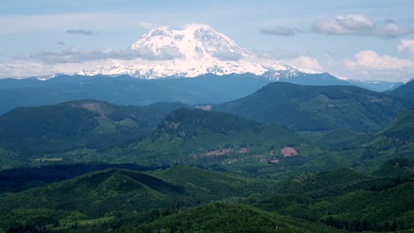 Snow-capped Mount Rainier rises above rolling green hills and forested ridges under a partly cloudy sky in Washington State.
