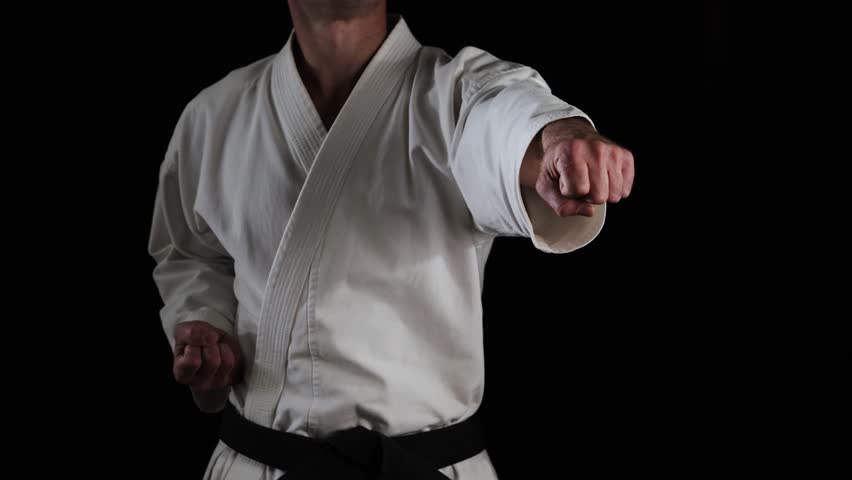 A male athlete with a black belt in karategi practices punches and blocks with his hands on a black background