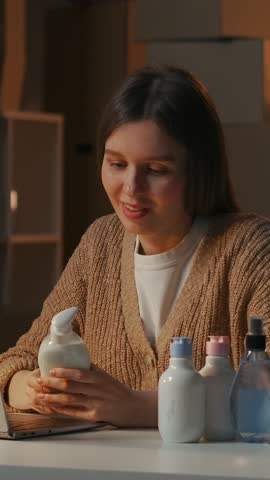 Young woman reviewing cosmetic skincare bottles at desk, examining product packaging and labels in warm indoor lighting while working on beauty routine content