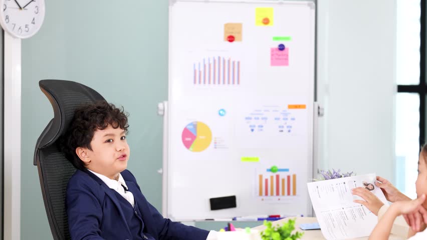 A young boy in a suit sits at a desk talking during a corporate meeting
