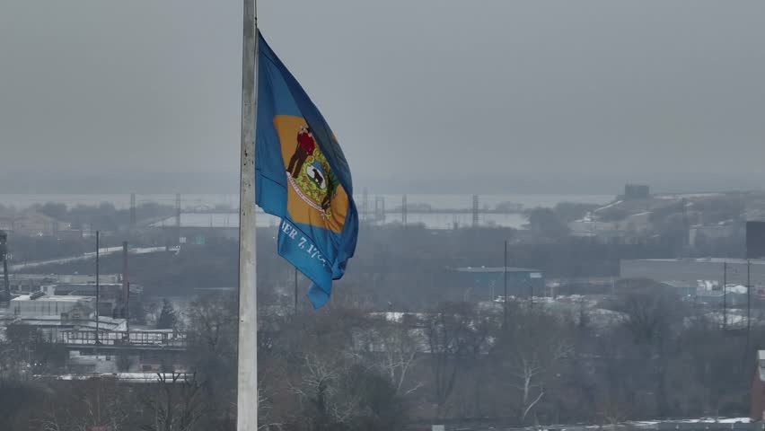 Close aerial drone view of Delaware state flag waving on a pole during cold winter weather with snowy landscape and distant city skyline visible through foggy atmosphere. Patriotic state in USA.