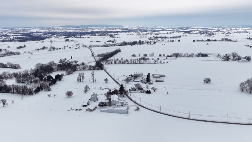 Wide aerial drone view of snowy countryside in Pennsylvania with farms, barns and rural roads crossing white winter fields. Quiet agricultural landscape stretches to distant horizon. Peaceful suburbia