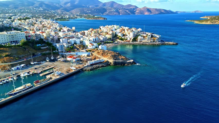 Drone aerial view of Agios Nikolaos harbor with white buildings along the coastline and blue sea on a sunny day.