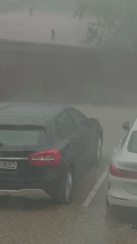 Vertical Screen: A strong hailstorm hits a city parking lot, with large hailstones falling and rain pouring down. Cars are left under the severe weather conditions as visibility drops.