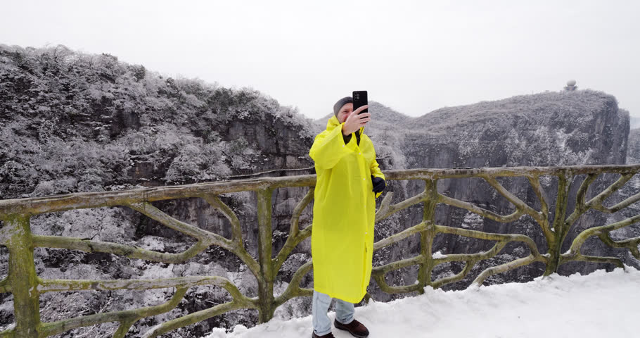 Tourist man in a bright yellow raincoat standing on a scenic viewpoint at cliff-side path and taking a selfie with a smartphone against a spectacular backdrop of snowy mountains in Tianmen Mountain National Forest Park. Fresh snow in late March after night cold storm create beautiful white appearance of bushes on mountain at background.