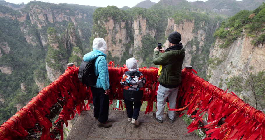 Tourist family with a child standing on viewpoint platform in Wulingyuan National Park, looking at the spectacular karst pillar landscape while the father is taking pictures with his smartphone. Red ribbons tied around railings on whole balcony, with long ribbons flying gently in wind, slow motion appearance