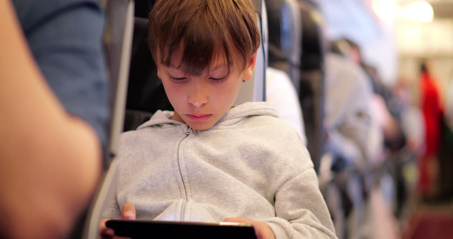 Handheld shot of young boy concentrating on a tablet while playing a videogame or watching a movie to stay entertained during a flight. He sits in an economy class aisle seat during a long-haul journey. Selective focus shot capturing the busy cabin atmosphere.