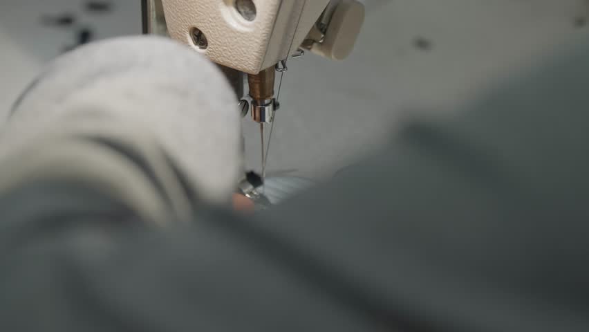 Close up industrial sewing machine needle stitching dark fabric while hands guide garment