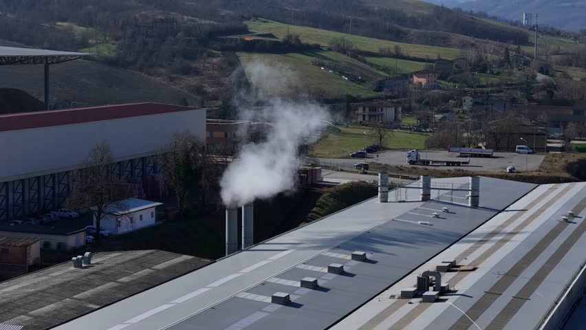 Factory facility with metal roofs and chimneys emitting white vapor into the air in an industrial production area, aerial telephoto drone shot.