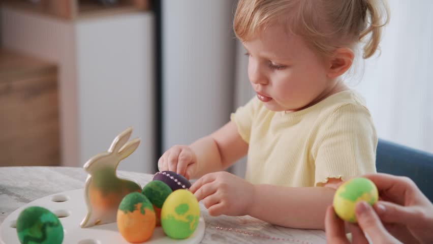 Little girl carefully paints Easter eggs with bright colors. She focuses intently, showing calm concentration. An adult gently holds another egg nearby. The scene captures joyful