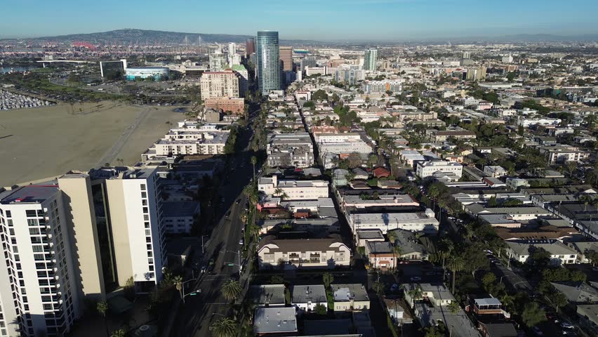 Coastal corridor view along Ocean Blvd near Alamitos Beach with downtown skyline, mid rise residences framing shoreline. Roof textures, palms, industrial port background in Long Beach, California. USA