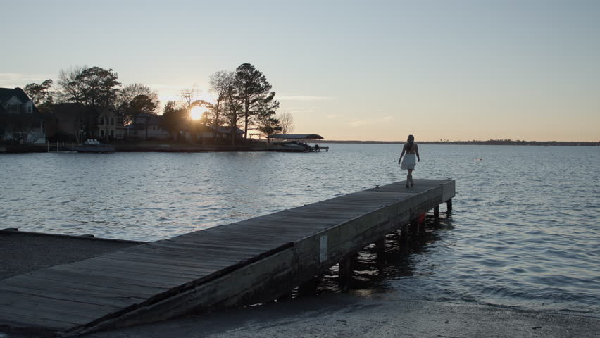 Wide camera shot of a beautiful blonde girl walking down a boat dock at sunset