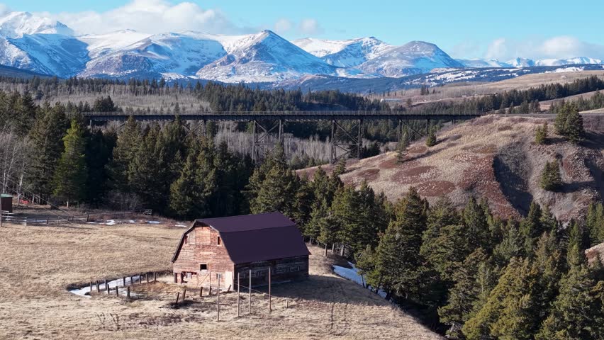 Drone footage shows a rustic barn standing on a grassy field in a rural valley with snow-capped mountains and coniferous trees near Whitefish, Montana, on a clear day.