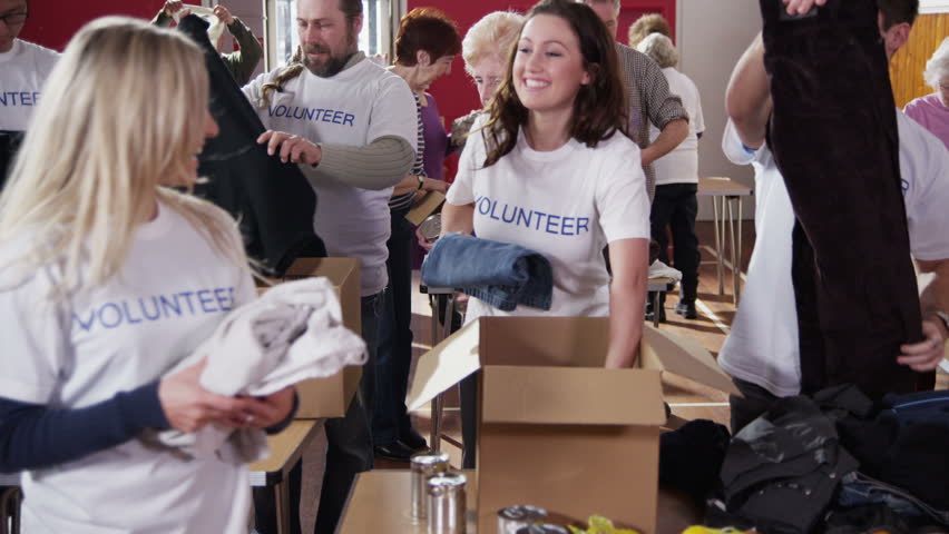 As charity workers and members of the community work together filling boxes with food and clothing, one female volunteer comes to the front of the shot and smiles directly at the camera. Slow motion. | Shutterstock HD Video #4027885