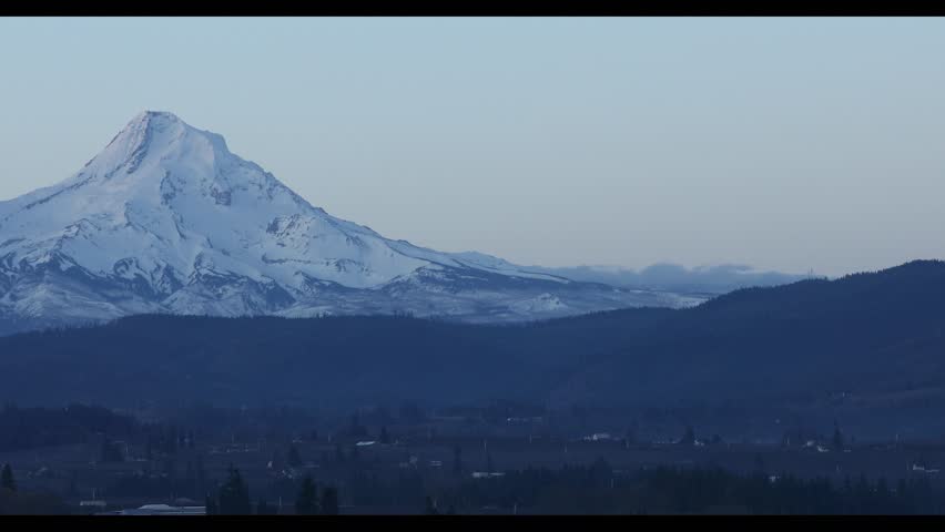 Slow panning close view of the snow covered summit of Mount Hood as seen from Hood River, Oregon, USA during an early winter morning just before sunrise. The iconic Cascade volcano rises above layered hills and the Columbia River Gorge region, creating a dramatic Pacific Northwest mountain landscape. Calm natural atmosphere with cold blue tones and winter conditions highlighting the rugged alpine peak and surrounding terrain. Scenic nature landscape footage ideal for travel media, environmental visuals, outdoor storytelling, presentation visuals and marketing content. Cinematic 4K mountain footage suitable for background motion and nature b roll.
