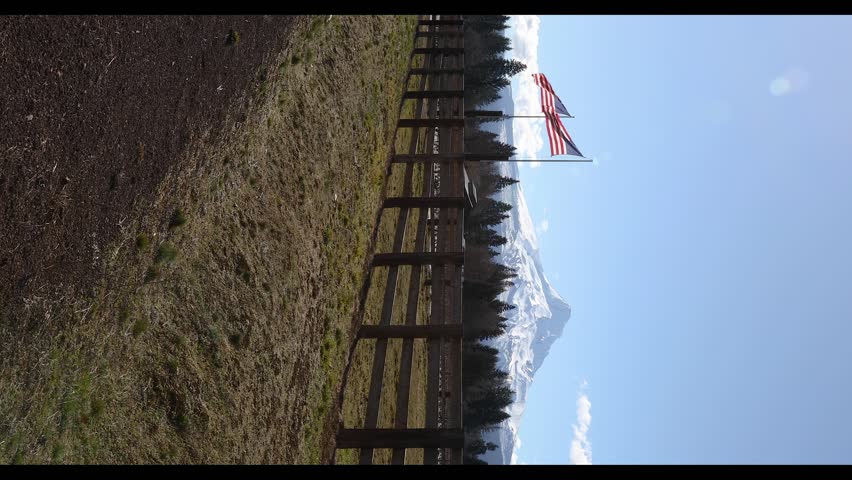 American flag waving in the wind with Mount Hood rising in the background in rural countryside near Hood River, Oregon, USA. Scenic Pacific Northwest landscape with snow covered mountain and evergreen forest under clear blue sky. Patriotic national symbol representing freedom, independence and pride in the United States. Ideal visual for Independence Day, July 4th themes, national identity, travel media, marketing projects and outdoor nature storytelling in cinematic 4K vertical footage with natural movement perfect for background visuals and b roll.