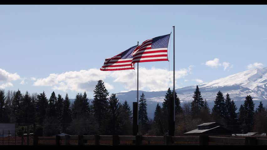 American flag waving in the wind with Mount Hood rising in the background in rural countryside near Hood River, Oregon, USA. Scenic Pacific Northwest landscape with snow covered mountain and evergreen forest under clear blue sky. Patriotic national symbol representing freedom, independence and pride in the United States. Ideal visual for Independence Day, July 4th themes, national identity, travel media, marketing projects and outdoor nature storytelling in cinematic 4K footage with natural movement perfect for background visuals and b roll.