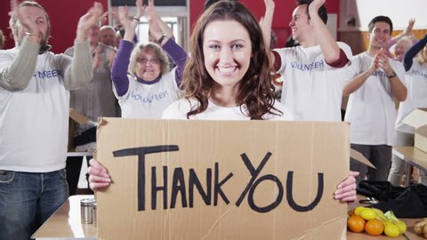 An attractive female charity volunteer holds up a "Thank You" sign and smiles at the camera as her fellow volunteers applaud and cheer in the background.