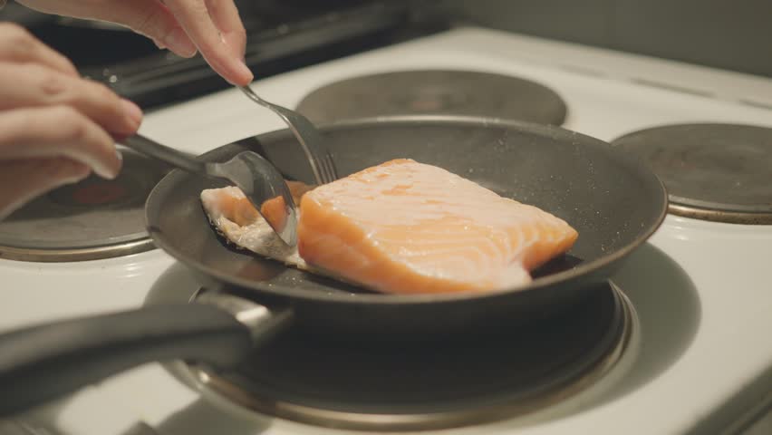 A close-up view of salmon being cooked in a frying pan, showcasing the sizzling moment. A hand is seen using a spatula to flip the fish, highlighting the cooking process in a kitchen environment.