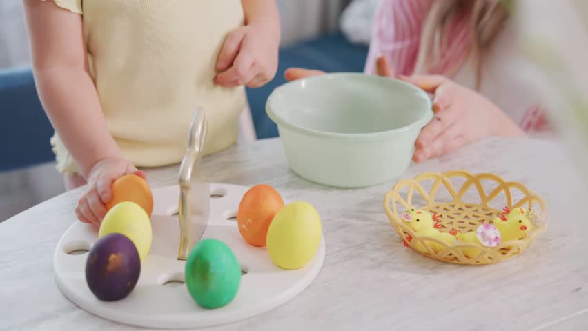 Children paint colorful Easter eggs at a table. Small hands carefully dip eggs in dye. Brightly colored eggs sit on a white plate. One child holds a basket of decorations. The scene feels joyful