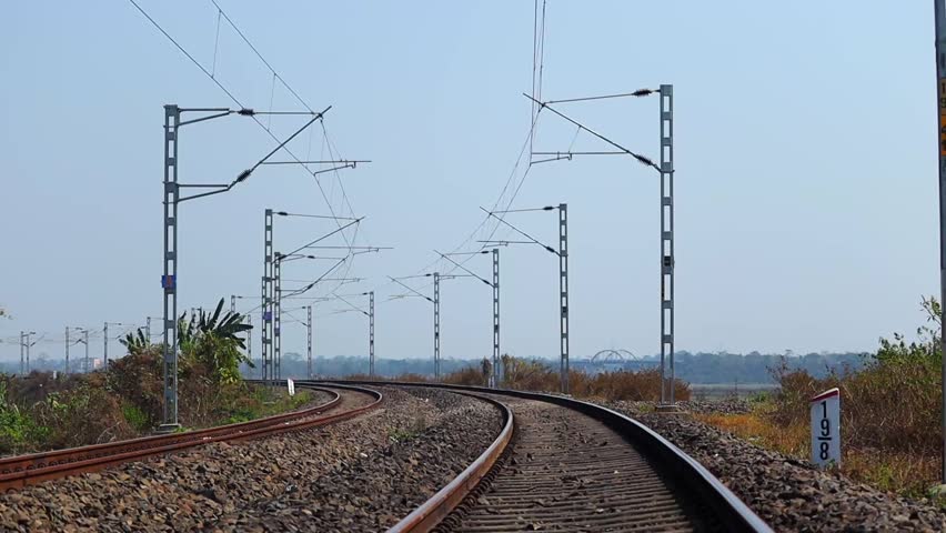 A railway track curving through a rural landscape with overhead electric railway poles and cables stretching into the distance under a clear blue sky. The scene highlights transportation infrastructure and railway connectivity across open countryside terrain.