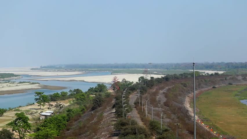 Scenic landscape showing a wide river with sandbanks, winding water channels, and a rural embankment road with trees and light poles under a clear sky.