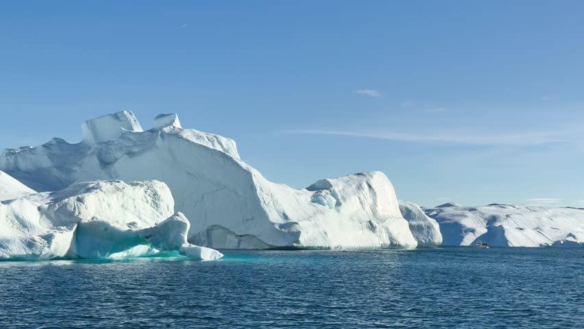 Large icebergs float in the water as the sun shines over Greenland. The clear blue sky contrasts with the white ice formations that fill the landscape. Boats move nearby while exploring.