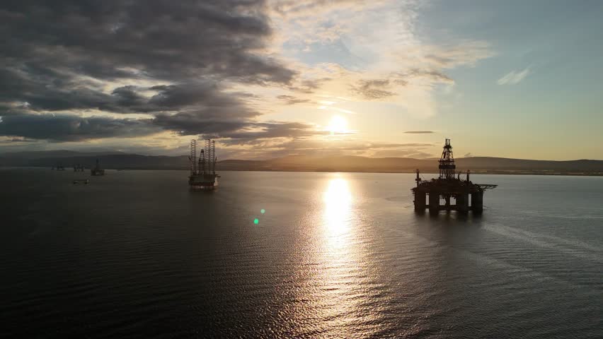 Aerial cinematic reverse shot moving away from decommissioned oil platforms anchored in Cromarty Firth, Scotland, during a dramatic golden hour sunset.