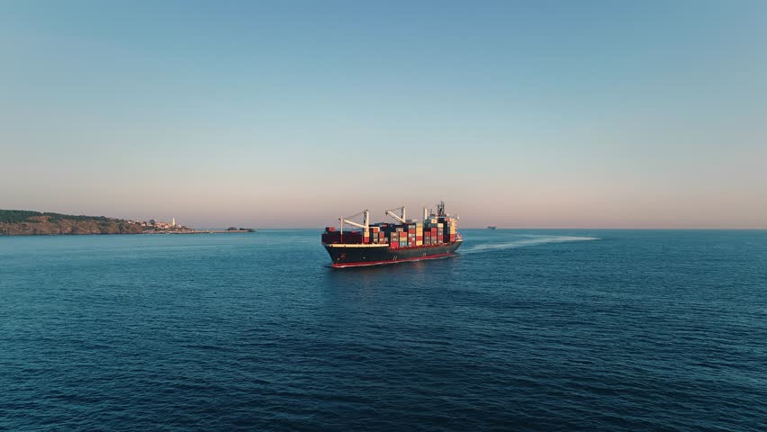 Fully loaded container ship underway on calm open sea at golden hour with seagull in flight, aerial drone wide shot with coastal silhouette in background