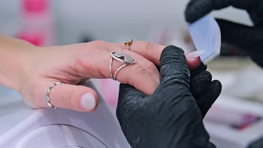 Salon manicure detail showing black gloved hands refining the edge of a long fingernail with an emery board while supporting the hand.