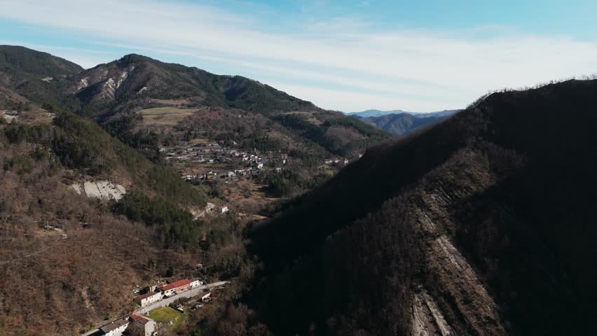 Aerial landscape of a narrow mountain valley with scattered village homes, wooded slopes and winding roads beneath a soft blue sky.