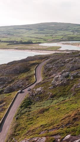Coastal road drive on a sunny day along the Wild Atlantic Way in Ireland, with cars traversing green hills and a peaceful bay.