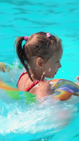 Happy little girl plays and laughs in blue pool with swimming ring. Child fun swims and splashes with rubber ring. Summer vacation holiday travel. Vertical shot