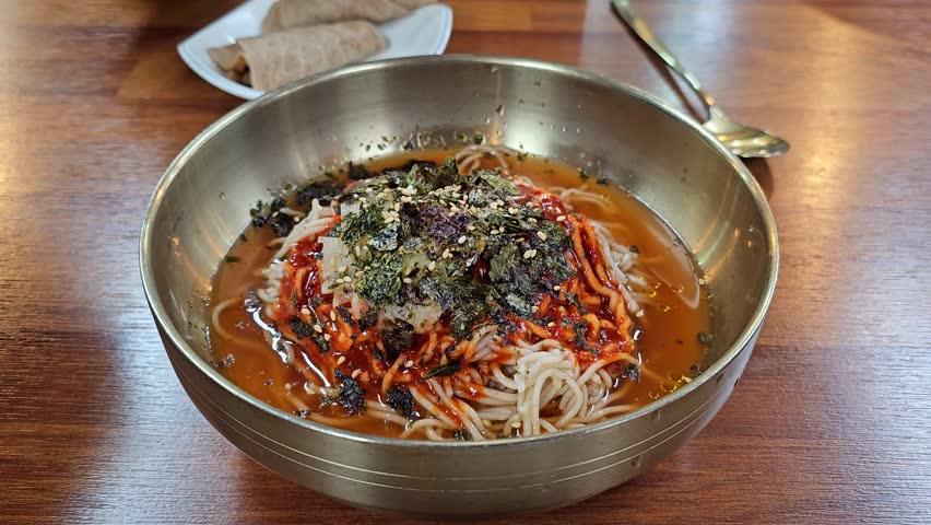 Chopsticks stir spicy red gochujang buckwheat noodles topped with dried seaweed and sesame seeds in traditional Korean steel bowl served with bossam wrap