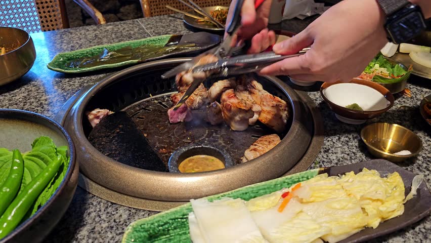 Hands use tongs to flip grilling samgyeopsal pork belly on Korean BBQ tabletop grill surrounded by fresh vegetables, dipping sauces and traditional side dishes