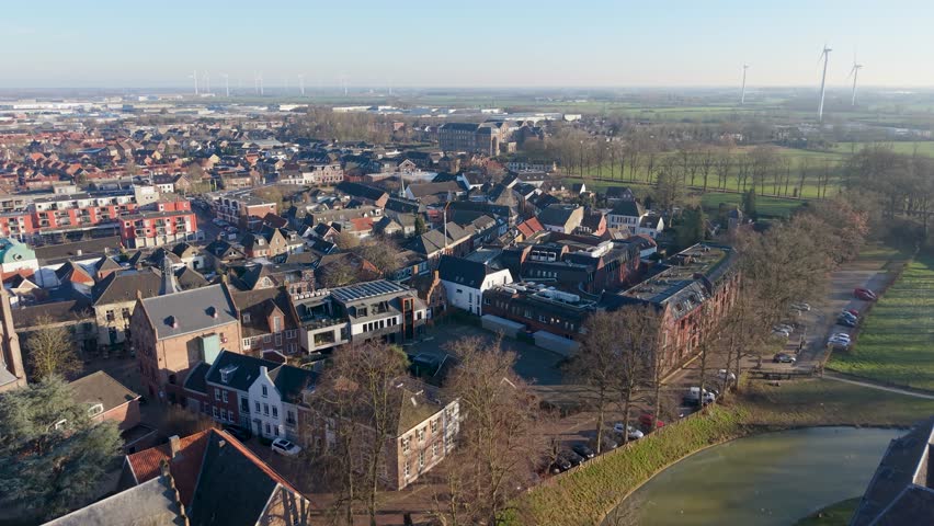 Aerial view of a scenic European town with historic brick architecture, a quiet canal, and a modern wind farm on the horizon under a clear blue sky. A perfect blend of heritage and green energy.