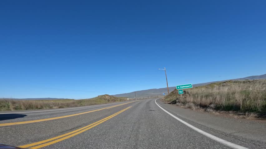 Driving POV along Highway 197 toward The Dalles Dam North Shore viewpoint in Washington State, near The Dalles, Oregon, USA in the Columbia River Gorge region. Scenic rural road through dry hills and open landscape on a sunny day with clear blue sky. Transportation route near the Columbia River connecting Oregon and Washington in the Pacific Northwest. Ideal footage for travel in USA, roads in USA, road trip concepts, transportation infrastructure, tourism visuals, logistics themes and driving perspective media. Cinematic 4K roadway footage captured by action camera suitable for background visuals and driving b roll.