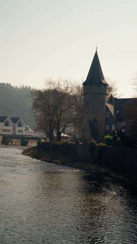Riverside Medieval Tower At Dawn, Soft Light Over Calm River, Leafless Trees Along Stone Embankment, Small Bridge And White Houses In Background, Misty Hills Beyond, Tranquil Atmosphere