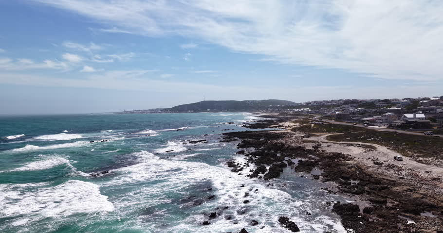 Drone aerial view of the rugged rocky shoreline and turquoise ocean waves along the coast near Struisbaai in the Western Cape, South Africa. Coastal homes and a seaside road run alongside the shoreline while waves break across the rocks along the scenic coastline near Cape Agulhas.