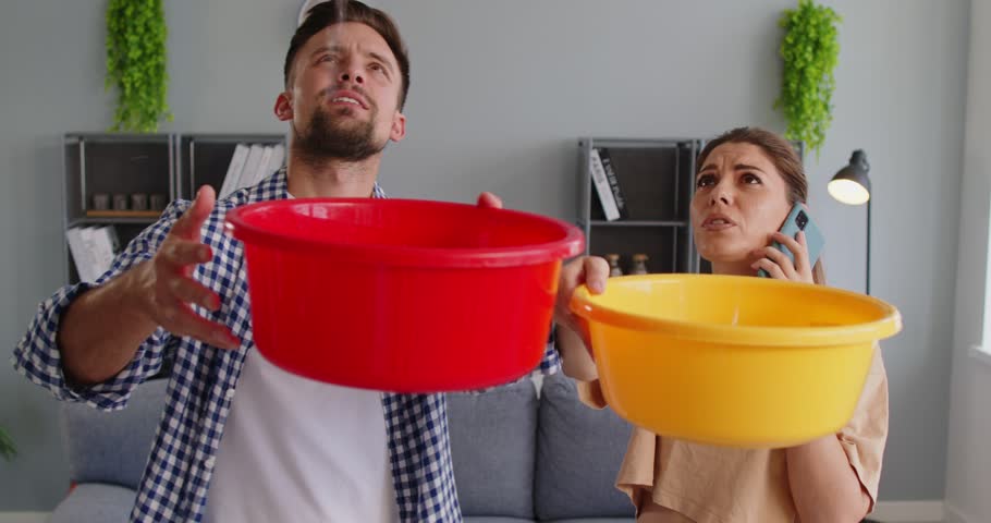 Couple holding buckets under ceiling leak. At home, they catch water falling from roof while calling for repair to reduce damage and stains. Leakage, home maintenance and emergency response.