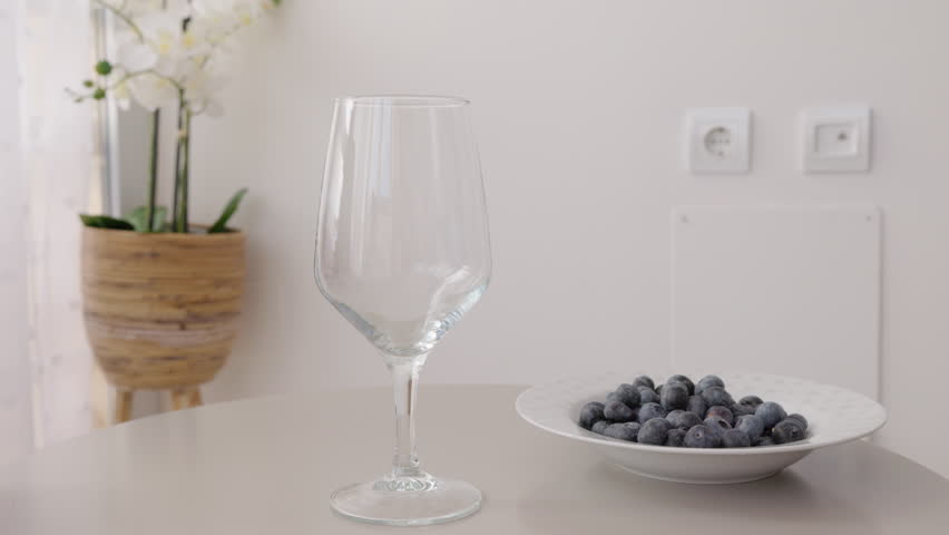 Hand pouring fresh orange juice into glass on kitchen table, blueberries on plate in bright modern home interior.