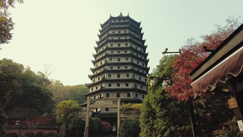 Hangzhou, China. A tall, multitiered pagoda with ornate architecture stands in a park setting, surrounded by trees and foliage. The sky is clear, and the sun casts a warm, golden hue over the scene.