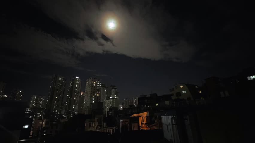 Full moon above residential city skyline with fast moving clouds across night sky, illuminated apartment buildings under dramatic moonlight creating atmospheric urban night scene.