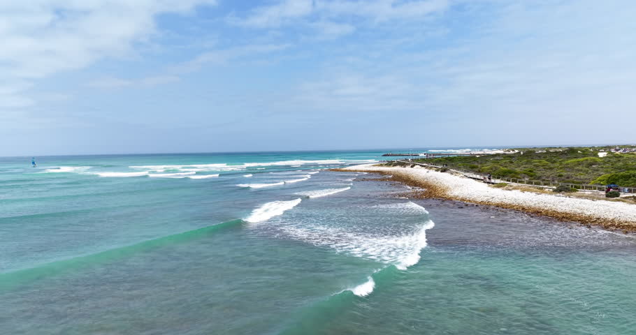 Beautiful coastal view of turquoise ocean waves rolling toward the rocky shoreline near the harbour in Struisbaai, Western Cape, South Africa. A wooden coastal boardwalk runs along the dunes while the calm bay and open sea stretch toward the horizon near Cape Agulhas, creating a peaceful seaside landscape.