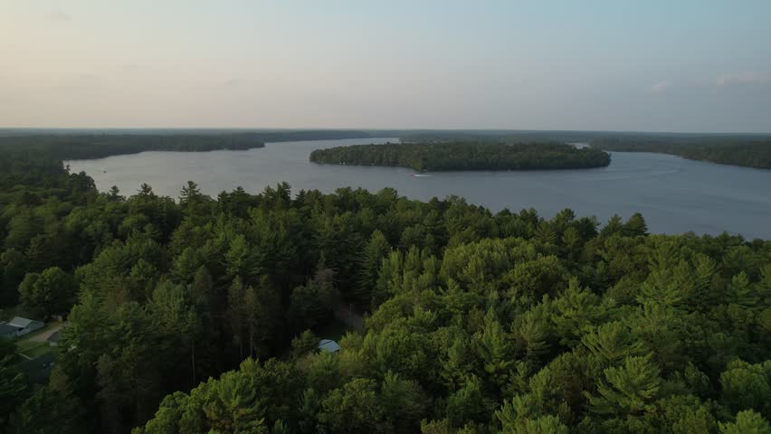 Aerial view of Eagle River Wisconsin in the Northwoods during summer.  Slow drone shot at golden hour over tree line, boat on lake in distance with island.
