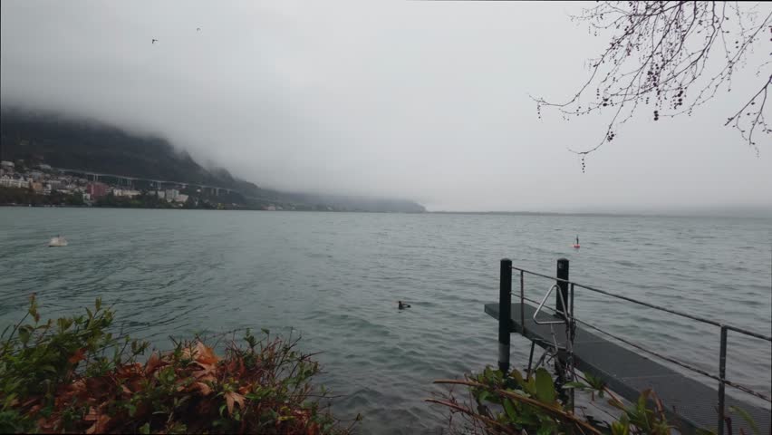 Foggy lakeshore view on Lake Geneva in Montreux, Switzerland with a small pier extending into the water. Mist covers the mountains while birds fly over the lake, creating a calm and atmospheric natural landscape.