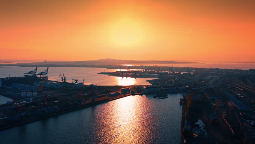 Vibrant sunset over maritime cargo port and harbor. Wide drone view of industrial cranes and port facilities with strong sun reflection.