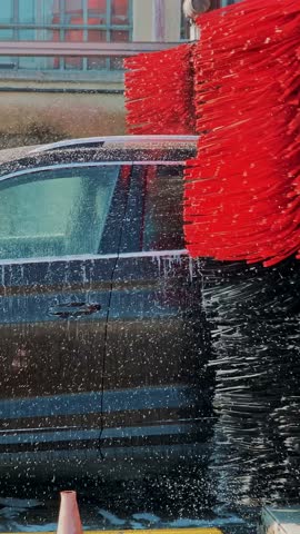 Dark SUV moving through automatic car wash station. Large red and black spinning brushes cleaning side doors with foam.