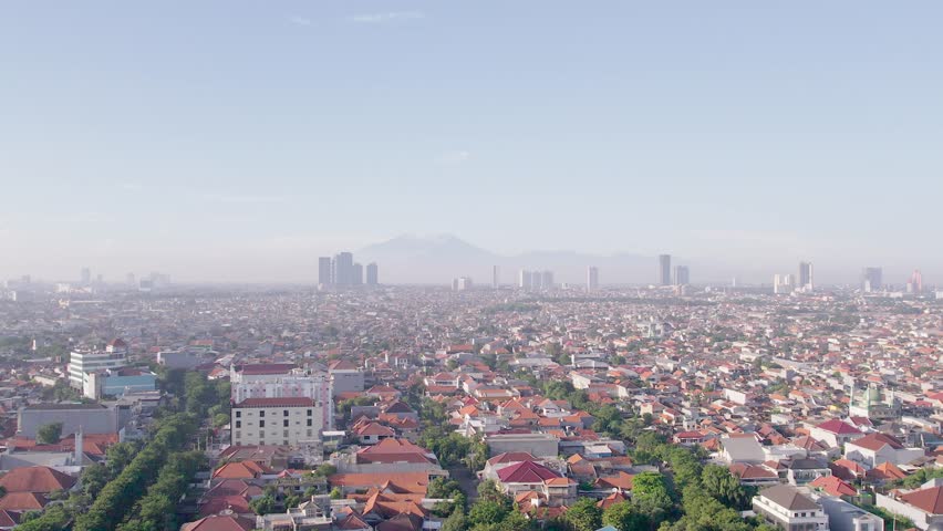 4K Aerial view of the dense urban cityscape of Surabaya, Indonesia, featuring a vast expanse of residential houses with orange-tiled roofs, modern high-rise buildings and a hazy mountain range